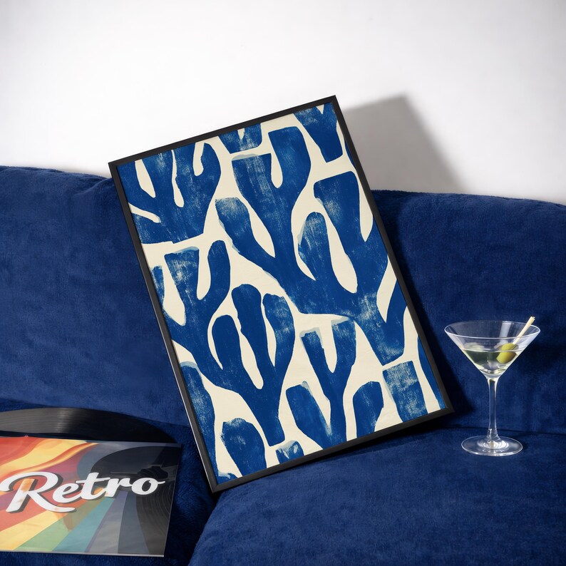 Blue abstract botanical poster in a white frame leaning against a white sofa, featuring deep navy leaf and coral-like organic shapes on a cream background, block print texture inspired by Matisse style cutout art. Modern coastal wall decor for living room or bedroom, ideal navy wall art print combining mid-century minimalist design with artisanal block printing texture — a striking indigo abstract statement piece for gallery walls and contemporary interiors.