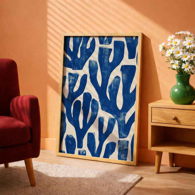 Blue abstract botanical poster in a white frame leaning against a white sofa, featuring deep navy leaf and coral-like organic shapes on a cream background, block print texture inspired by Matisse style cutout art. Modern coastal wall decor for living room or bedroom, ideal navy wall art print combining mid-century minimalist design with artisanal block printing texture — a striking indigo abstract statement piece for gallery walls and contemporary interiors.