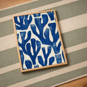 Blue abstract botanical poster in a white frame leaning against a white sofa, featuring deep navy leaf and coral-like organic shapes on a cream background, block print texture inspired by Matisse style cutout art. Modern coastal wall decor for living room or bedroom, ideal navy wall art print combining mid-century minimalist design with artisanal block printing texture — a striking indigo abstract statement piece for gallery walls and contemporary interiors.