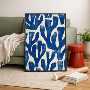 Blue abstract botanical poster in a white frame leaning against a white sofa, featuring deep navy leaf and coral-like organic shapes on a cream background, block print texture inspired by Matisse style cutout art. Modern coastal wall decor for living room or bedroom, ideal navy wall art print combining mid-century minimalist design with artisanal block printing texture — a striking indigo abstract statement piece for gallery walls and contemporary interiors.