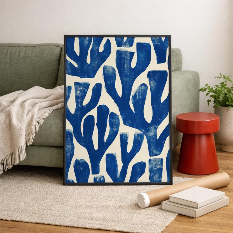 Blue abstract botanical poster in a white frame leaning against a white sofa, featuring deep navy leaf and coral-like organic shapes on a cream background, block print texture inspired by Matisse style cutout art. Modern coastal wall decor for living room or bedroom, ideal navy wall art print combining mid-century minimalist design with artisanal block printing texture — a striking indigo abstract statement piece for gallery walls and contemporary interiors.