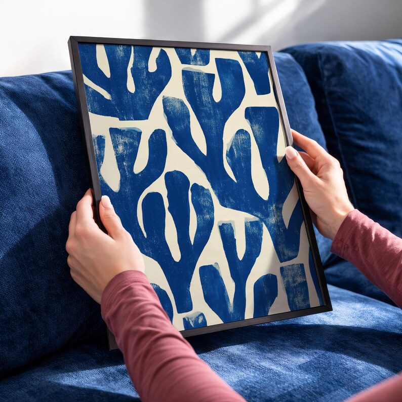 Blue abstract botanical poster in a white frame leaning against a white sofa, featuring deep navy leaf and coral-like organic shapes on a cream background, block print texture inspired by Matisse style cutout art. Modern coastal wall decor for living room or bedroom, ideal navy wall art print combining mid-century minimalist design with artisanal block printing texture — a striking indigo abstract statement piece for gallery walls and contemporary interiors.