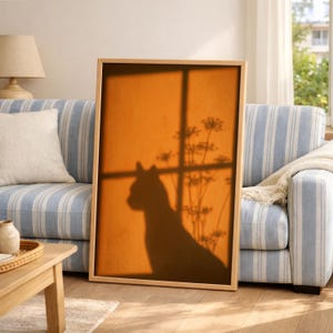 Cat shadow poster featuring a minimalist orange silhouette of a cat sitting by a sunlit window, with soft plant shadows on a textured amber wall. Moody aesthetic wall art perfect for bedrooms, reading nooks, or hallways. Warm sunset-toned cat silhouette print, a unique gift for cat lovers who enjoy dreamy, atmospheric wall decor rather than literal pet portraits.