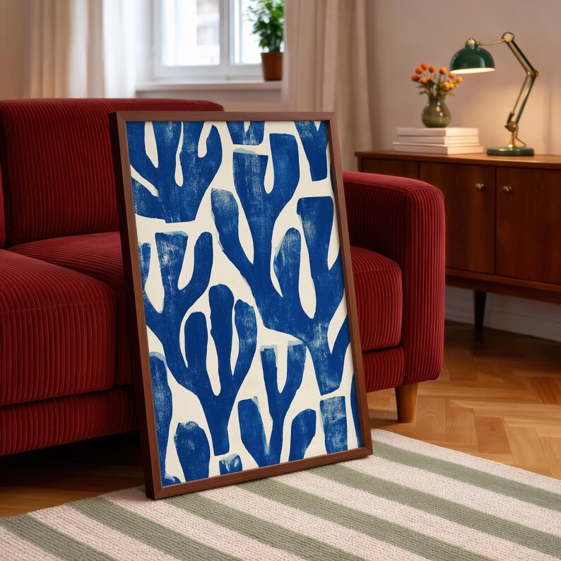 Blue abstract botanical poster in a white frame leaning against a white sofa, featuring deep navy leaf and coral-like organic shapes on a cream background, block print texture inspired by Matisse style cutout art. Modern coastal wall decor for living room or bedroom, ideal navy wall art print combining mid-century minimalist design with artisanal block printing texture — a striking indigo abstract statement piece for gallery walls and contemporary interiors.