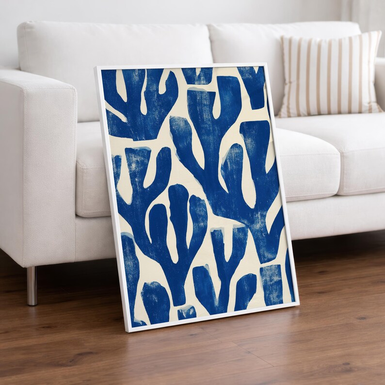Blue abstract botanical poster in a white frame leaning against a white sofa, featuring deep navy leaf and coral-like organic shapes on a cream background, block print texture inspired by Matisse style cutout art. Modern coastal wall decor for living room or bedroom, ideal navy wall art print combining mid-century minimalist design with artisanal block printing texture — a striking indigo abstract statement piece for gallery walls and contemporary interiors.