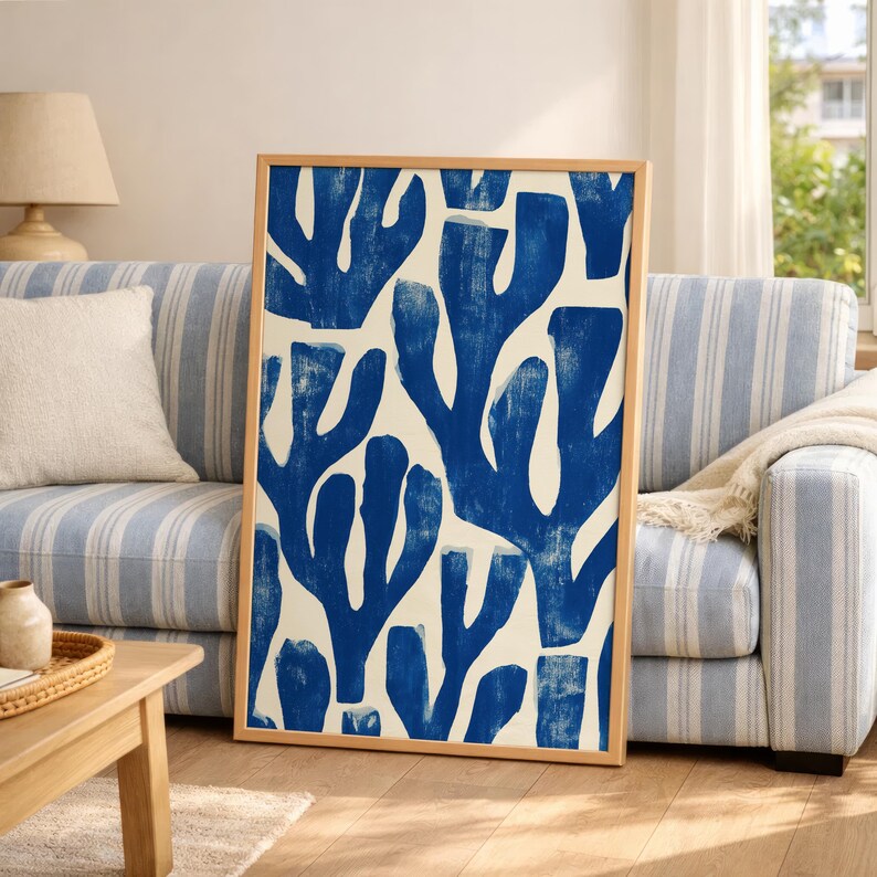 Blue abstract botanical poster in a white frame leaning against a white sofa, featuring deep navy leaf and coral-like organic shapes on a cream background, block print texture inspired by Matisse style cutout art. Modern coastal wall decor for living room or bedroom, ideal navy wall art print combining mid-century minimalist design with artisanal block printing texture — a striking indigo abstract statement piece for gallery walls and contemporary interiors.