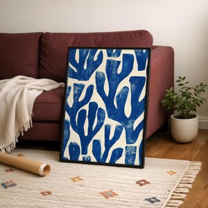 Blue abstract botanical poster in a white frame leaning against a white sofa, featuring deep navy leaf and coral-like organic shapes on a cream background, block print texture inspired by Matisse style cutout art. Modern coastal wall decor for living room or bedroom, ideal navy wall art print combining mid-century minimalist design with artisanal block printing texture — a striking indigo abstract statement piece for gallery walls and contemporary interiors.