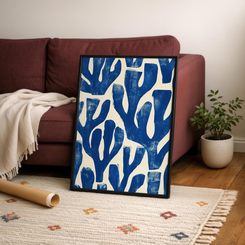 Blue abstract botanical poster in a white frame leaning against a white sofa, featuring deep navy leaf and coral-like organic shapes on a cream background, block print texture inspired by Matisse style cutout art. Modern coastal wall decor for living room or bedroom, ideal navy wall art print combining mid-century minimalist design with artisanal block printing texture — a striking indigo abstract statement piece for gallery walls and contemporary interiors.