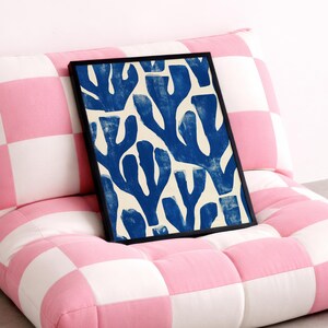 Blue abstract botanical poster in a white frame leaning against a white sofa, featuring deep navy leaf and coral-like organic shapes on a cream background, block print texture inspired by Matisse style cutout art. Modern coastal wall decor for living room or bedroom, ideal navy wall art print combining mid-century minimalist design with artisanal block printing texture — a striking indigo abstract statement piece for gallery walls and contemporary interiors.