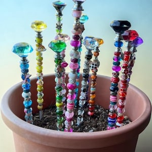 May include: A collection of decorative plant stakes featuring metal rods adorned with various colorful glass and plastic beads, topped with large faceted crystal-like gems in shades of blue, yellow, green, pink, orange, black, red, and purple.