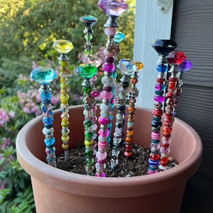 May include: A terracotta pot filled with soil and topped with colorful beaded garden stakes. The stakes feature various colored beads, including pink, blue, green, yellow, and red.