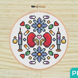 Puede incluir: Un aro de bordado de punto de cruz blanco con un diseño colorido que presenta dos riñones rojos, flores azules, hojas verdes, corazones rosas y jeringas. El diseño está rodeado de píldoras y otros símbolos médicos.