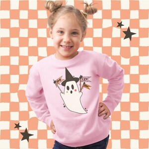 Puede incluir: Una sudadera rosa claro con un fantasma de dibujos animados que lleva un sombrero de bruja y sostiene banderas con las palabras "Yay" y "Ekk". El fantasma est&aacute; rodeado de estrellas. El fondo es un patr&oacute;n de cuadros.