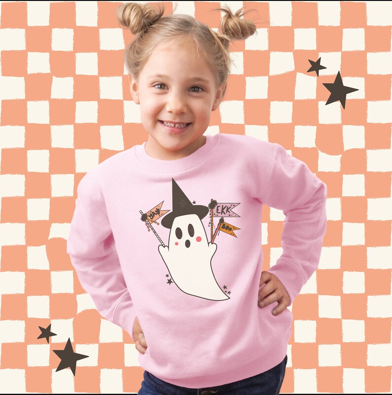 Puede incluir: Una sudadera rosa claro con un fantasma de dibujos animados que lleva un sombrero de bruja y sostiene banderas con las palabras "Yay" y "Ekk". El fantasma est&aacute; rodeado de estrellas. El fondo es un patr&oacute;n de cuadros.