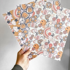 Op de afbeelding: Een set van drie gepatroneerde cadeaupapier met een retro jaren 70-stijl. De papieren bevatten regenbogen, vredestekens, bloemen, sterren en smiley-gezichten. Een papier heeft de tekst "Girls Rule" die overal wordt herhaald.