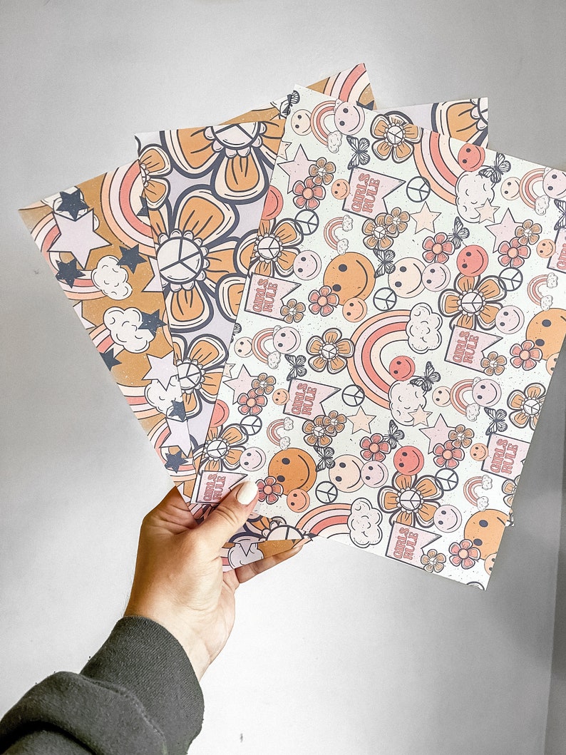 Op de afbeelding: Een set van drie gepatroneerde cadeaupapier met een retro jaren 70-stijl. De papieren bevatten regenbogen, vredestekens, bloemen, sterren en smiley-gezichten. Een papier heeft de tekst "Girls Rule" die overal wordt herhaald.