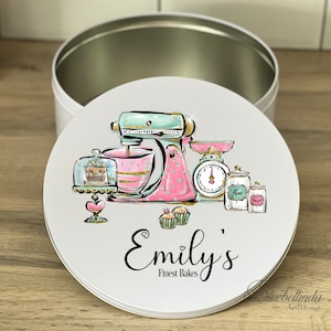 May include: White round tin featuring a pink and green illustration of a stand mixer, a cake stand with a cake, a scale, and two jars. The text "Emily's Finest Bakes" is printed below the illustration.