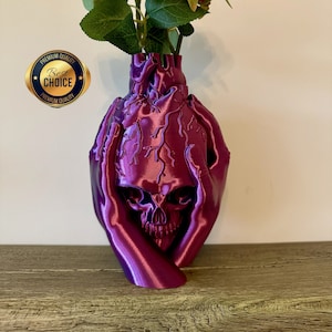 May include: A purple skull vase with two red roses. The skull is surrounded by two hands, and the vase has a textured surface. The vase is labelled "Premium Quality Best Choice Premium Quality".