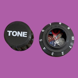 May include: A black knob with the word "TONE" printed on it and a black numbered dial with a clear window revealing guitar picks inside.