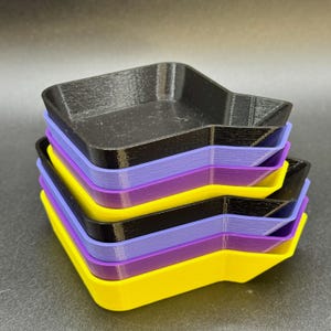 May include: A stack of colourful, square-shaped trays in black, purple, yellow, and lavender. The trays have a unique, angular design and are stacked neatly. The trays are made of a solid material.