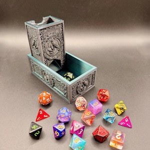May include: A dark grey, rectangular dice tower with an ornate, embossed raven design on the front panel. The tower features an attached tray base containing one die, while a collection of multi-coloured, translucent, and solid polyhedral dice are scattered in front of the structure.