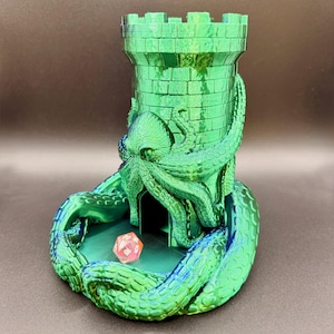 May include: A green, metallic dice tower in the shape of a castle, with an octopus design. A pink, multi-sided die sits inside the tower's base. The tower is designed for tabletop gaming.