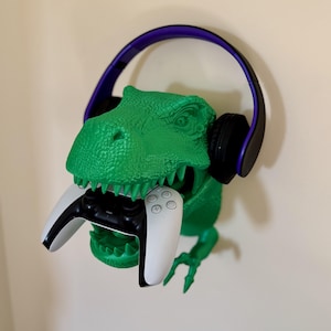 May include: A green dinosaur head wall mount with a purple and black headset and a white and black video game controller in its mouth.