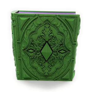 May include: A green decorative book with detailed embossed patterns and a central diamond-shaped feature. The book's edges are a lighter green, and the pages are purple. The cover has ornate floral and geometric designs.