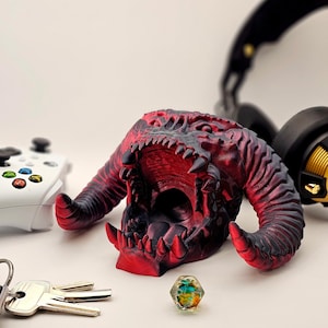 May include: A red and black 3D printed dragon head with an open mouth, used as a headphone stand. The dragon head is sitting on a white surface with a gaming controller and a multi-coloured dice.