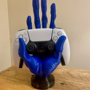 May include: A blue 3D printed hand holding a white PlayStation 5 controller. The hand is posed with the fingers extended and the palm facing forward. The controller is resting in the palm of the hand. The hand is mounted on a black base.