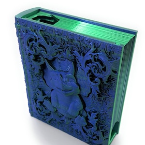 May include: A decorative book in blue and green, featuring detailed carvings of floral patterns and a creature. The edges and top of the book are green, with the cover displaying a blue relief design. The background is white.