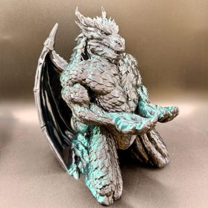 May include: A metallic dark grey dragon figurine in a kneeling position with its hands extended forward. The creature features detailed scales, sharp horns, and large wings folded behind its back, with subtle teal highlights on the body.