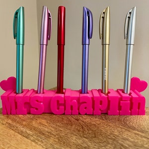 May include: Pink plastic pen holder with the name "Mrs. Chaplin" in raised letters. The holder has six slots for pens and two heart-shaped cutouts. Six pens are in the holder, including a teal, pink, red, purple, gold, and silver pen.