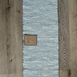 Peut inclure: Un tapis rectangulaire tissé avec un motif à rayures bleues et blanches. Le tapis a un bord effiloché et une petite étiquette attachée. Le tapis est posé sur un sol en bois.