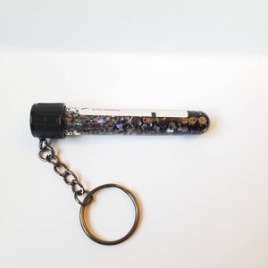 Op de afbeelding: Een zwarte reageerbuis sleutelhanger gevuld met iriserende glitter. De reageerbuis heeft een zwarte schroefdop en een wit label met de tekst "2 No Additive".