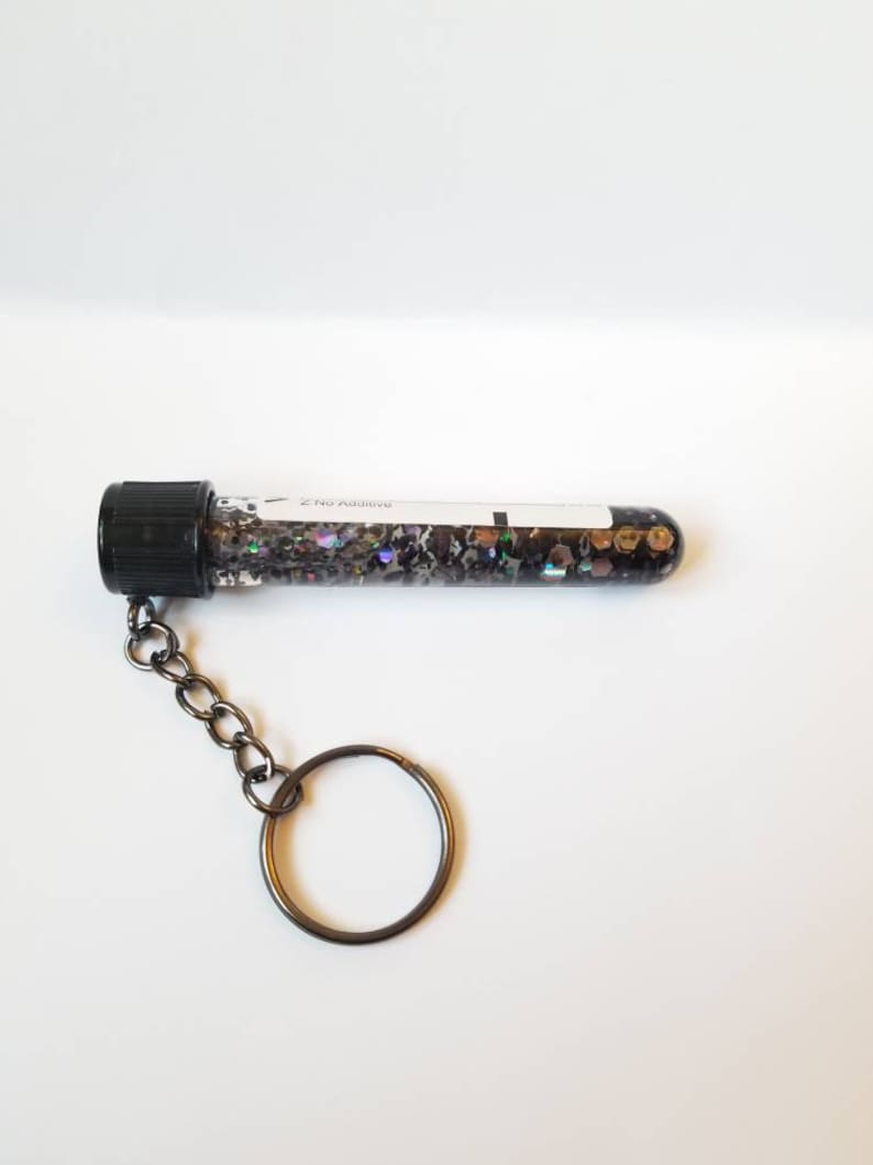 Op de afbeelding: Een zwarte reageerbuis sleutelhanger gevuld met iriserende glitter. De reageerbuis heeft een zwarte schroefdop en een wit label met de tekst "2 No Additive".