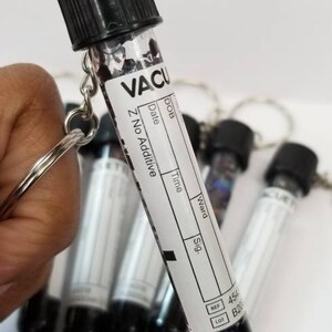 Op de afbeelding: Een klein, helder glazen flesje met een zwarte dop en een wit label met de tekst "VACU Z No Additive". Het flesje is bevestigd aan een zilveren sleutelhanger. Het flesje is gevuld met zwarte glitter.
