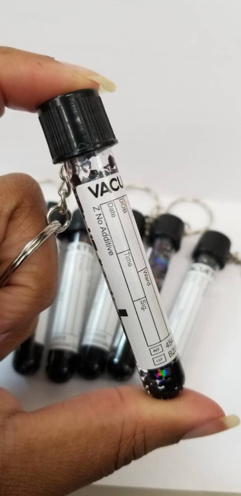 Op de afbeelding: Een klein, helder glazen flesje met een zwarte dop en een wit label met de tekst "VACU Z No Additive". Het flesje is bevestigd aan een zilveren sleutelhanger. Het flesje is gevuld met zwarte glitter.