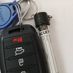 Op de afbeelding: Een zwarte autosleutel met vier knoppen en een rood lampje. De sleutel is bevestigd aan een sleutelhanger met een blauwe sleutelkaart en een klein flesje met het label "ACUET" met een wit label en zwarte tekst.