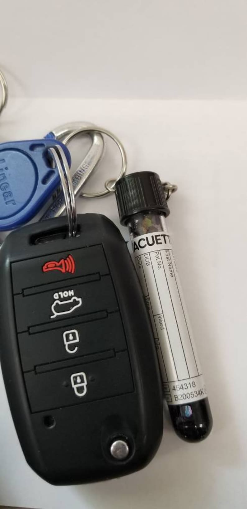 Op de afbeelding: Een zwarte autosleutel met vier knoppen en een rood lampje. De sleutel is bevestigd aan een sleutelhanger met een blauwe sleutelkaart en een klein flesje met het label "ACUET" met een wit label en zwarte tekst.