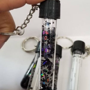Op de afbeelding: Een sleutelhanger in de vorm van een flesje gevuld met zwarte en iriserende glitter, met een zwarte dop en een zilveren ketting.
