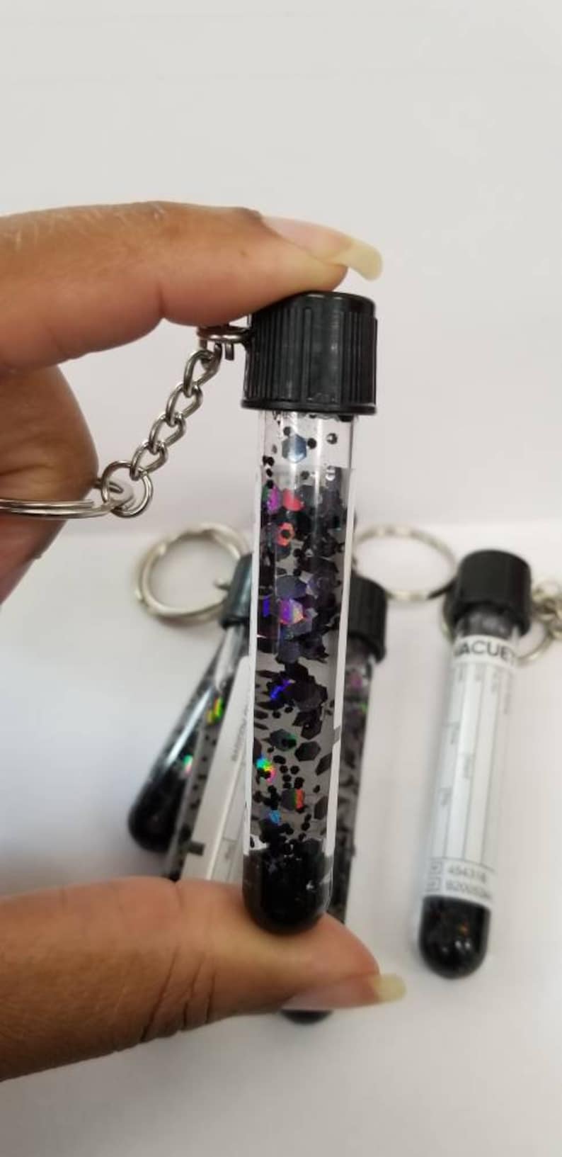 Op de afbeelding: Een sleutelhanger in de vorm van een flesje gevuld met zwarte en iriserende glitter, met een zwarte dop en een zilveren ketting.