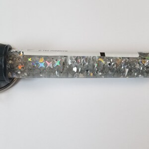 Op de afbeelding: Een transparante glazen buis gevuld met zilveren glitter en regenboogkleurige sterren, bevestigd aan een zwarte dop en een zilveren sleutelhanger. De buis heeft een wit label met de tekst "Rainbow 2 Nu" erop gedrukt.