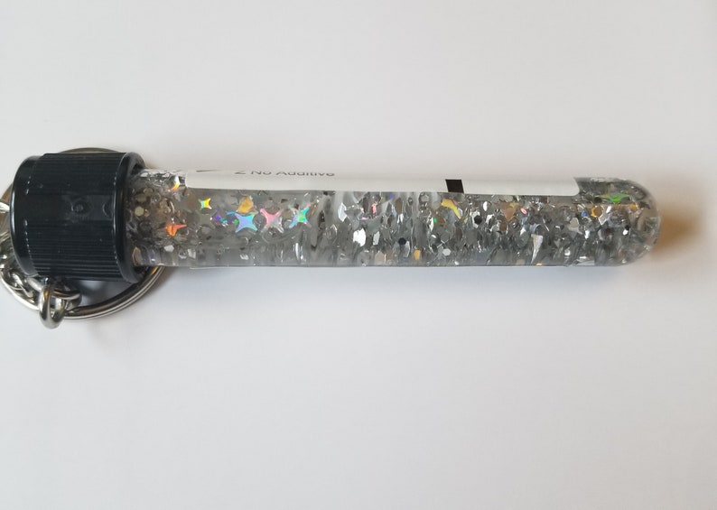 Op de afbeelding: Een transparante glazen buis gevuld met zilveren glitter en regenboogkleurige sterren, bevestigd aan een zwarte dop en een zilveren sleutelhanger. De buis heeft een wit label met de tekst "Rainbow 2 Nu" erop gedrukt.