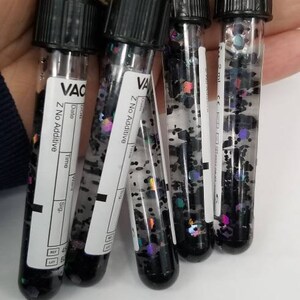 Op de afbeelding: Vijf kleine, doorzichtige glazen flesjes met zwarte glitter en zwarte dopjes, bevestigd aan een zilveren ketting. De flesjes zijn gelabeld met "VAC" en "Z No Additive".