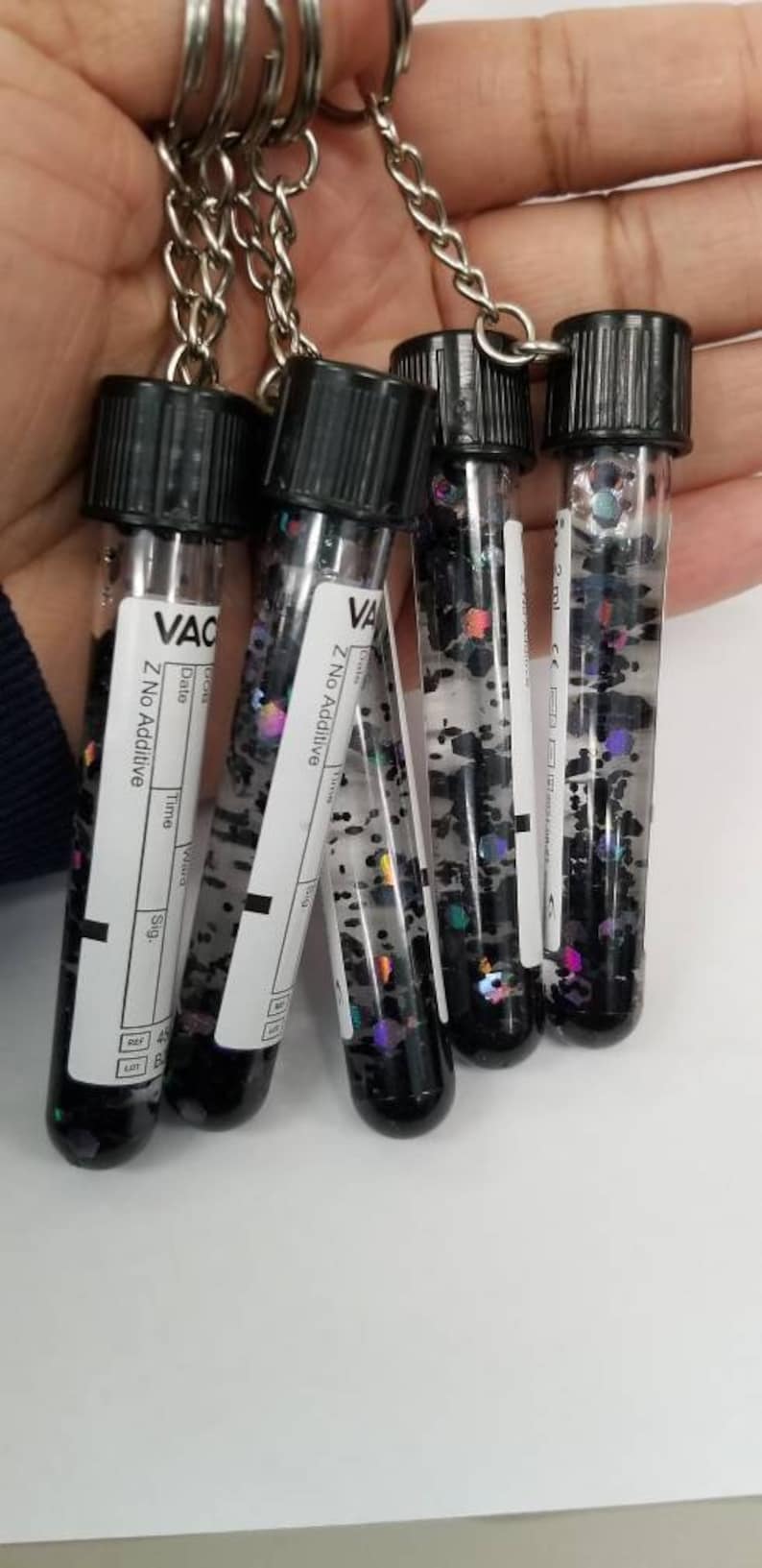 Op de afbeelding: Vijf kleine, doorzichtige glazen flesjes met zwarte glitter en zwarte dopjes, bevestigd aan een zilveren ketting. De flesjes zijn gelabeld met "VAC" en "Z No Additive".
