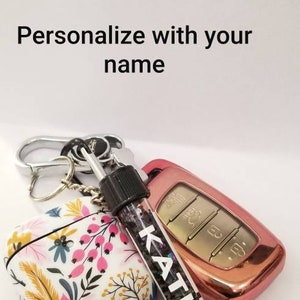 Op de afbeelding: Een sleutelhanger met een AirPod-hoesje met bloemenprint, een ros&eacute;gouden autosleutelhanger en een doorzichtig buisje gevuld met glitter waarop "KATIE" staat.