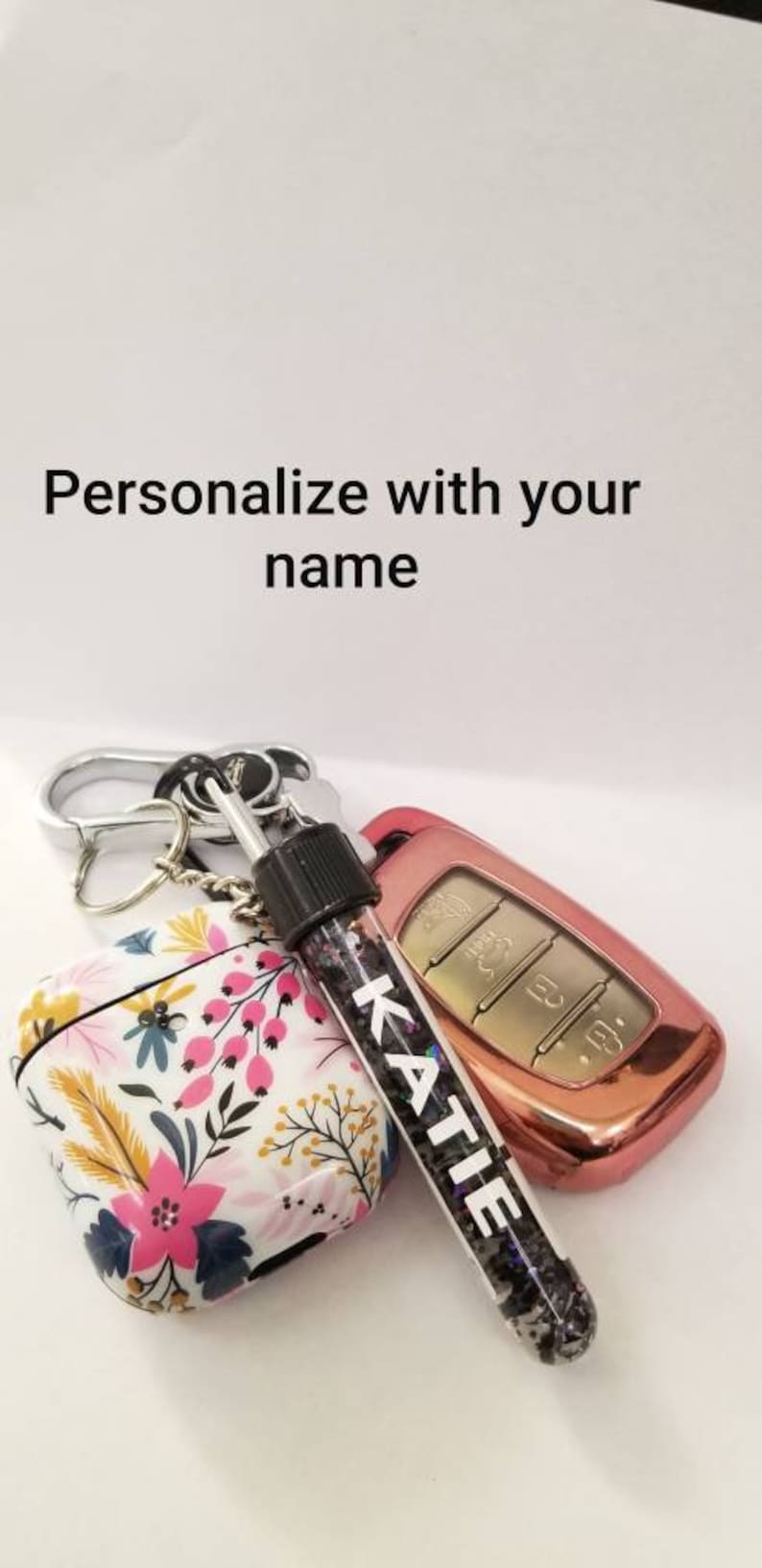 Op de afbeelding: Een sleutelhanger met een AirPod-hoesje met bloemenprint, een ros&eacute;gouden autosleutelhanger en een doorzichtig buisje gevuld met glitter waarop "KATIE" staat.
