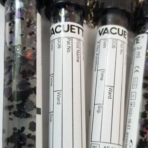 Op de afbeelding: Vier zwarte en witte flesjes met sleutelhangers. Elk flesje heeft een wit etiket met de tekst "VACUETTE" en velden voor "Achternaam", "Voornaam", "Pat.-nr.", "Geboortedatum", "Datum", "Tijd", "Afdeling" en "Handtekening". De flesjes zijn gevuld met kleurrijk glitter. Op de bodem van de flesjes staat de tekst "454318 B200534K" en "454318 B200534".