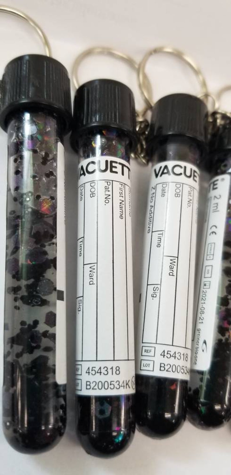 Op de afbeelding: Vier zwarte en witte flesjes met sleutelhangers. Elk flesje heeft een wit etiket met de tekst "VACUETTE" en velden voor "Achternaam", "Voornaam", "Pat.-nr.", "Geboortedatum", "Datum", "Tijd", "Afdeling" en "Handtekening". De flesjes zijn gevuld met kleurrijk glitter. Op de bodem van de flesjes staat de tekst "454318 B200534K" en "454318 B200534".